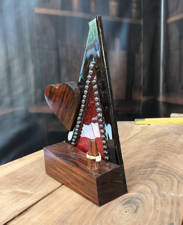 Bold and captivating, Crimson Covenant is a handcrafted ring holder that unites the natural beauty of exotic woods with radiant artistic expression. The base and sculpted heart are carved from richly grained, all-natural Cocobolo wood—renowned for its warmth and elegance. The vertical frame, made of striking Wenge wood, encloses a vibrant red stained glass panel infused with glitter and sealed beneath a glass-like epoxy finish. A ring post, also Cocobolo, awaits the symbol of a cherished union.