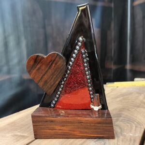 Bold and captivating, Crimson Covenant is a handcrafted ring holder that unites the natural beauty of exotic woods with radiant artistic expression. The base and sculpted heart are carved from richly grained, all-natural Cocobolo wood—renowned for its warmth and elegance. The vertical frame, made of striking Wenge wood, encloses a vibrant red stained glass panel infused with glitter and sealed beneath a glass-like epoxy finish. A ring post, also Cocobolo, awaits the symbol of a cherished union.