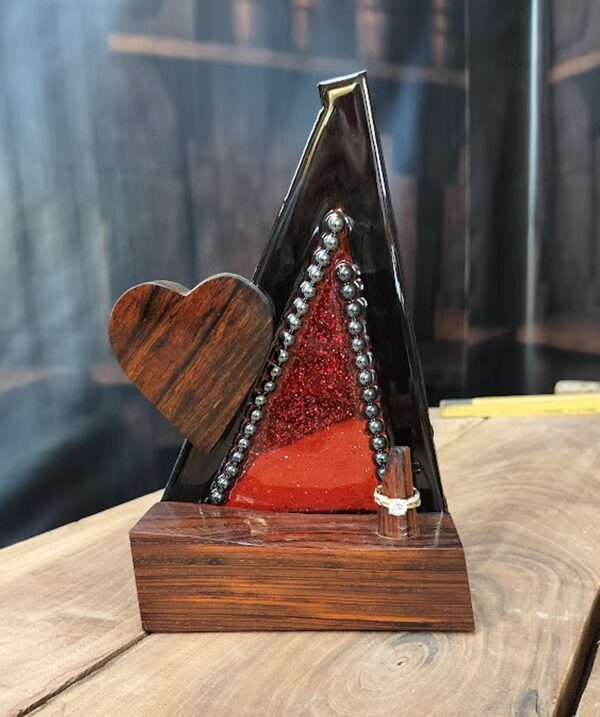 Bold and captivating, Crimson Covenant is a handcrafted ring holder that unites the natural beauty of exotic woods with radiant artistic expression. The base and sculpted heart are carved from richly grained, all-natural Cocobolo wood—renowned for its warmth and elegance. The vertical frame, made of striking Wenge wood, encloses a vibrant red stained glass panel infused with glitter and sealed beneath a glass-like epoxy finish. A ring post, also Cocobolo, awaits the symbol of a cherished union.