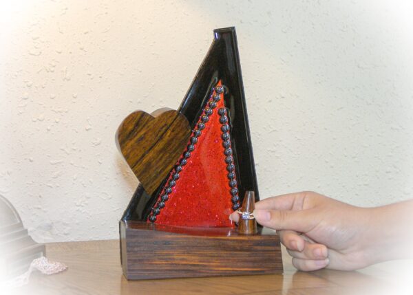 Bold and captivating, Crimson Covenant is a handcrafted ring holder that unites the natural beauty of exotic woods with radiant artistic expression. The base and sculpted heart are carved from richly grained, all-natural Cocobolo wood—renowned for its warmth and elegance. The vertical frame, made of striking Wenge wood, encloses a vibrant red stained glass panel infused with glitter and sealed beneath a glass-like epoxy finish. A ring post, also Cocobolo, awaits the symbol of a cherished union.