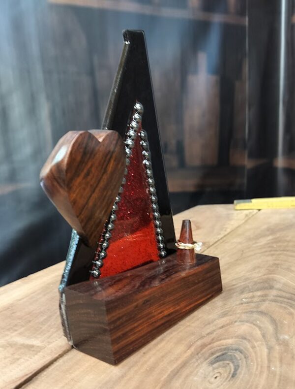 Bold and captivating, Crimson Covenant is a handcrafted ring holder that unites the natural beauty of exotic woods with radiant artistic expression. The base and sculpted heart are carved from richly grained, all-natural Cocobolo wood—renowned for its warmth and elegance. The vertical frame, made of striking Wenge wood, encloses a vibrant red stained glass panel infused with glitter and sealed beneath a glass-like epoxy finish. A ring post, also Cocobolo, awaits the symbol of a cherished union.
